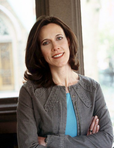 Author Megan McCafferty, who has shoulder-length brown hair, smiles in front of a lighted window with her arms crossed, wearing a teal-colored top and a grey jacket.