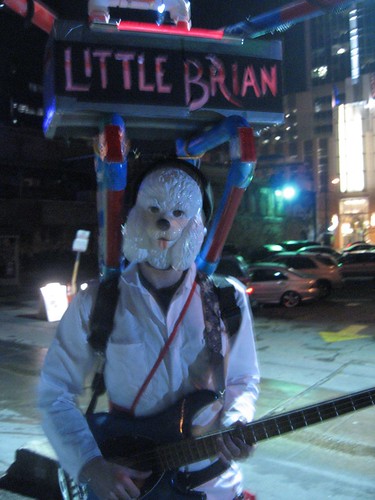 A man in a dog mask playing bass with a sign above that says Little Brian