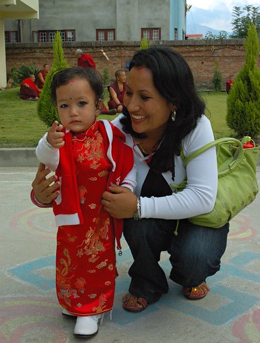 Hey there! Tibetan / Nepalese beauty in red gown, white cowboy boots, child with her mama, standing on a colored chalk Infinity Knot - auspicious symbol, Tharlam Monastery Courtyard,  Boudha, Kathmandu, Nepal by Wonderlane