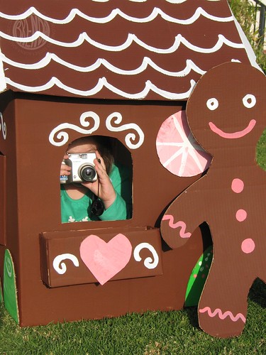 child inside a large cardboard gingerbread house with a camera in her hand
