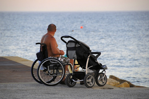 man in wheelchair sitting at jetty edge looking to the ocean. Next to him is a pram.