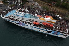 Photo du navire principal accosté avec une foule énorme pour célébrer son départ vers Gaza. Très long navire blanc, à plusieurs étages, avec les drapeaux de plusieurs mouvements sociaux.