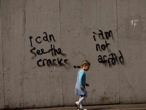 girlchild skipping on the sidewalk -- behind her, on a wall, the graffiti reads, "I can see the cracks -- I am not afraid."