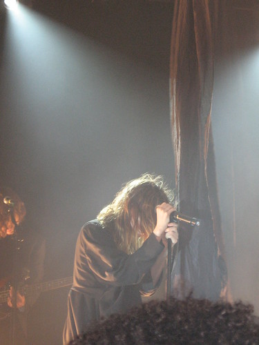 Lykke Li in front of a hanging black drape, holding the microphone with both hands with her head down, her face not visible behind her hair.