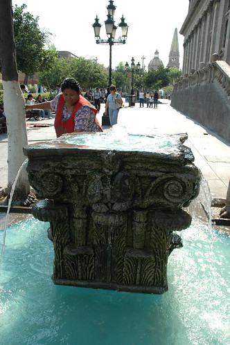 A Mexican woman playing with the water of a classic stone fountain on the site of founding, Cathedral in background, ornate black lamps, Downtown Guadalajara, Jalisco, Mexico by Wonderlane