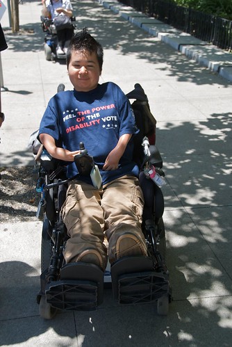 A person in a powerchair wearing a shirt that says 'feel the power of the disability vote.' Photo taken at a protest in California.