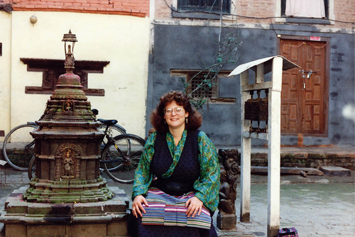 Linda Lane in a courtyard with a Buddhist shrine, prayer wheel, and prayer wheels, bicycles, window, wood door, wearing a dark colored Tibetan Chuba, green silk blouse, and striped apron, in Kathmandu, Nepal, in 1990 by Wonderlane