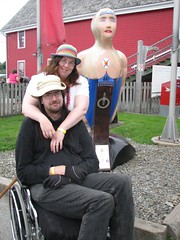 A white woman (Anna), sunburned and tired looking, stands behind a white man (Don), also sunburned and tired looking, in a manual wheelchair.  They are posed in front of a wooden figurehead of a mermaid
