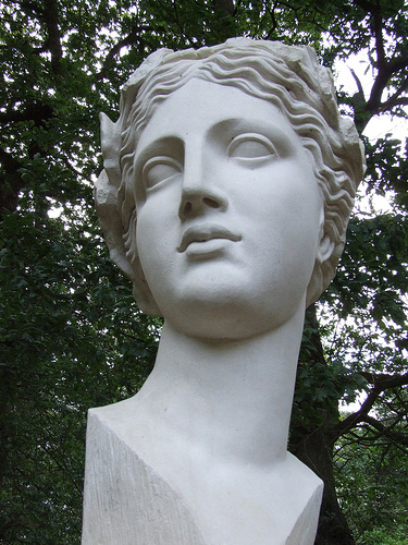 Close-up of a white bust of Sappho titled "Xth Muse" by Ian Hamilton Finlay. It is mostly a smooth, nondescript face but has textured strands of short hair. Evening sun shines through a treetop in the background.