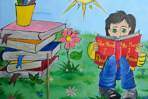 graffiti of a small green-eyed child reading a book, with a stack of books next to her; the title of the book reads, "the more I read/the more I know"