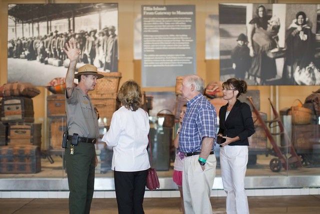 Sarah Palin stands with 3 family members inside the Ellis Island museum. A tour guide faces them and raises his hand, gesturing away from the back wall, which displays large photographs of immigrants from the early 20th century and many old valises and suitcases.