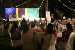 People sitting around dinner tables in front of a stage on the beach
