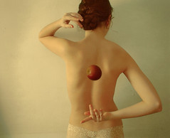 woman facing away from the camera.  She is caught in the middle of dropping an apple behind her back, both arms curved behind her.