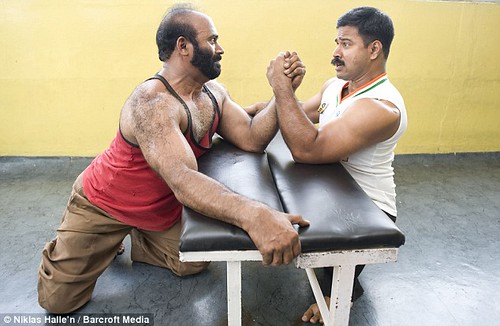Joby Matthew and an unidentified man arm-wrestle on a weight bench. The unidentified man, who has a beard and fully developed legs, grips the far side of the weight bench.  Both men are grimacing and neither appears to be winning.