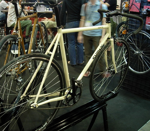 Rear, right side view of a tan Surly Cross Check bike, on a display stand, in a room with people standing around
