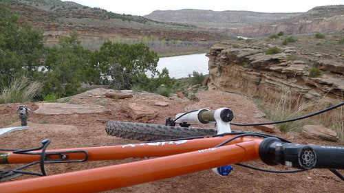 An orange Surly Troll bike laying on it's right side, on a cliff ledge, with a river canyon below in the background
