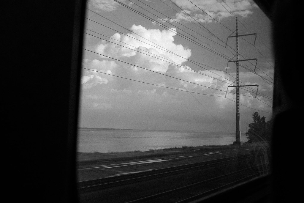 Coastal inlet beneath towering thunderheads flanked by utility towers, seen from a train window.