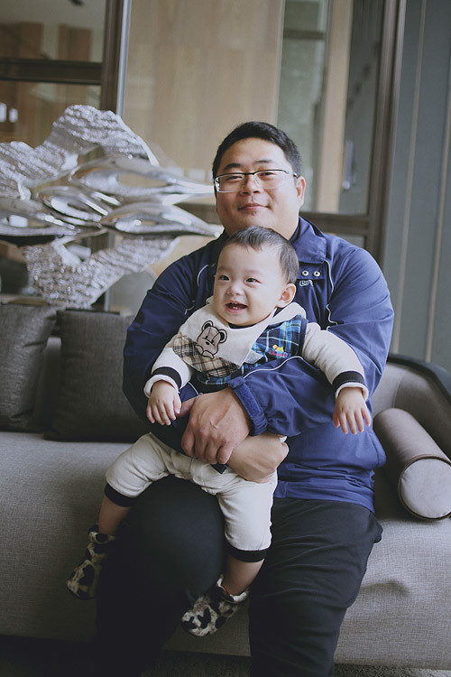 爸爸把兒子抱在腿上，坐在窗邊沙發上合照。